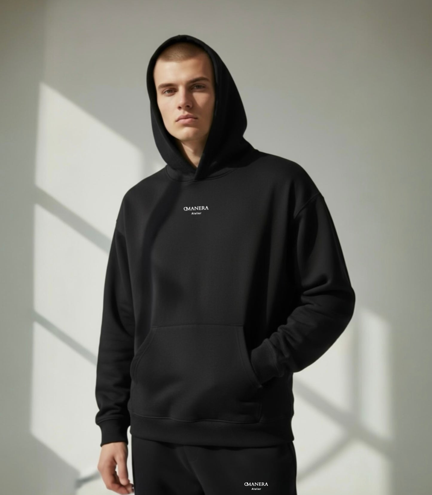 Junger Mann trägt schwarzen DMANERA Atelier Hoodie und Jogginghose, minimalistischer Look, Tageslicht