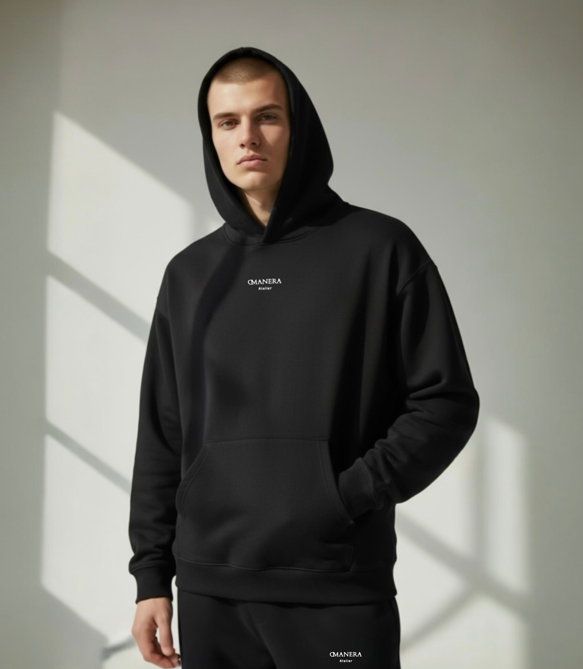 Junger Mann trägt schwarzen DMANERA Atelier Hoodie und Jogginghose, minimalistischer Look, Tageslicht